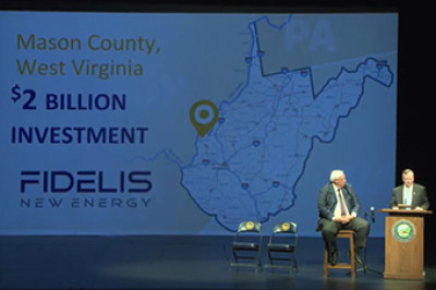 two men on a stage in front of a screen displaying text and a map of the state of West Virginia