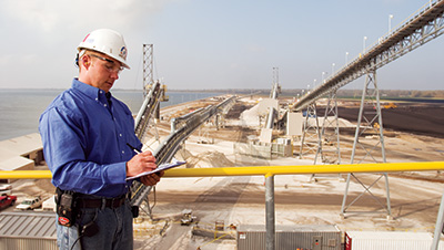 Plant operator on elevated platform writing notes on clipboard wearing hard hat and looking over exterior view of power plant